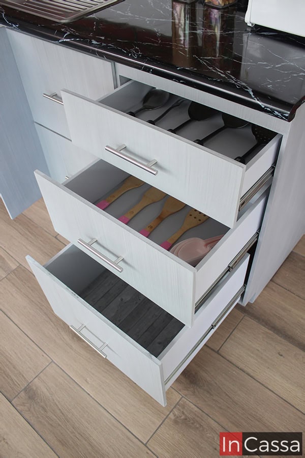 Se nos muestran tres de los cajones del módulo inferior, los cuales son presentados abiertos dejando ver su interior y estructura completa, los cuales mantienen el mismo acabado cenizo claro que puede apreciarse en toda la cocina. También se aprecian mejor detalles como son: los rieles metálicos y las jaladeras de barra T integrados a cada cajón.
