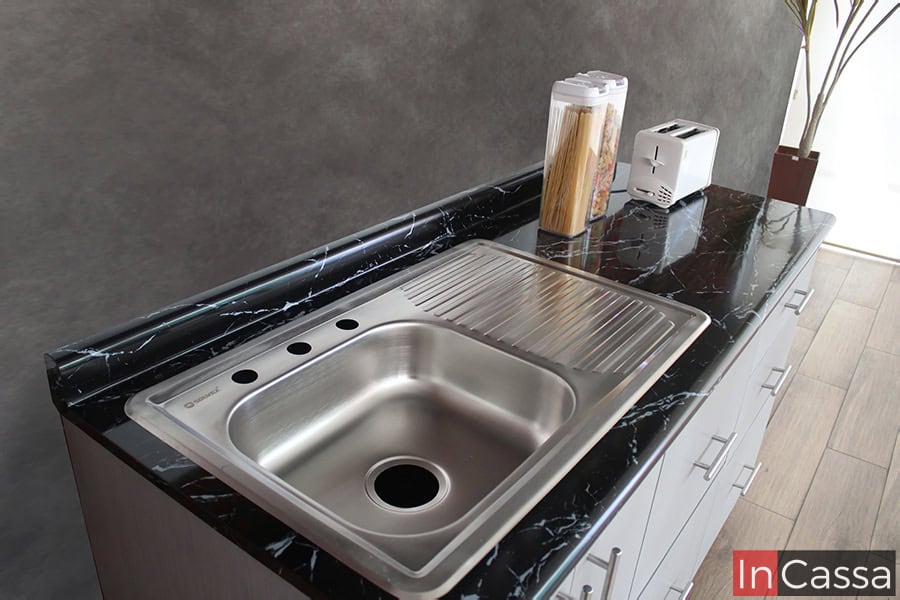 La foto de enfoca en la cubierta de formaica del módulo inferior de la cocina, la cuál tiene una acabado estilo mármol negro y cuenta con una tarja de acero inoxidable con escurridor incorporada.