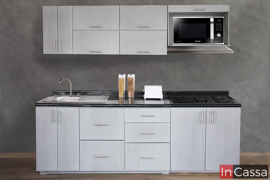 Una toma frontal de la cocina en la que es posible apreciar todos sus módulos, en la que resalta su acabado gris cenizo claro, su cubierta de formaica en estilo mármol negro y su espacio para microondas en el mueble sobre campana.