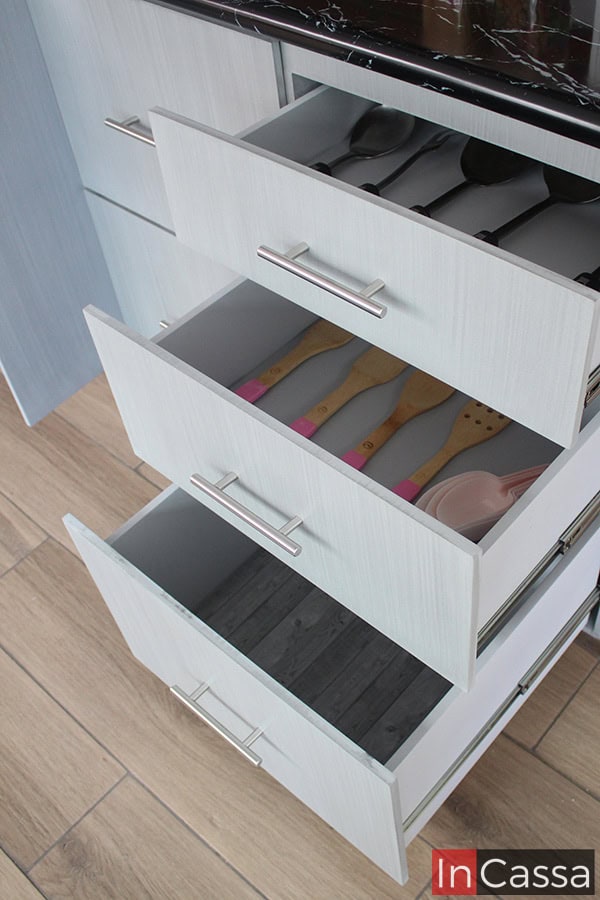 Una vista cercana de tres de los cajones del módulo inferior de la cocina. Gracias a que son mostrados abiertos, podemos ver la estructura completa de cada cajón, misma que mantiene completamente el mismo acabado cenizo claro que puede verse en el resto de la cocina. También podemos apreciar pequeños detalles como son los rieles metálicos y las jaladeras de barra T integrados a cada uno de los cajones.