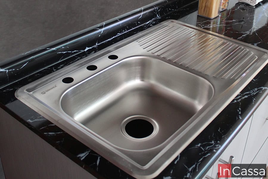Una foto de la tarja de acero inoxidable incluida en la cocina, está cuenta con una tina sencilla y un escurridor. Por la cercanía de la toma, también tenemos una mejor vista de la cubierta de formaica de estilo mármol negro.