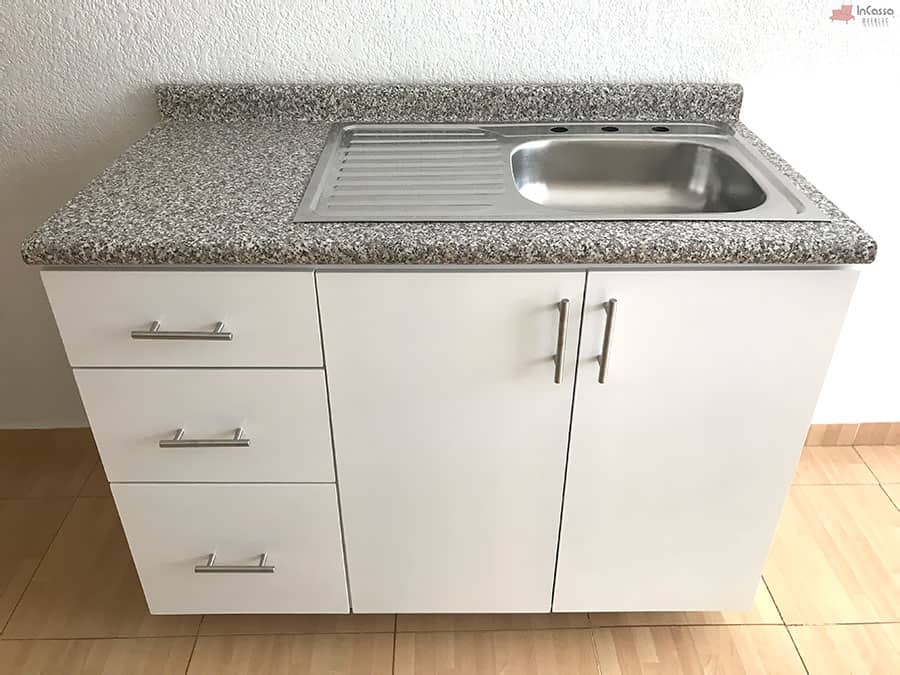 La imagen muestra una toma frontal del módulo inferior de la Cocina Integral NEVADA de 2 m en color blanco, destacando sus tres amplios cajones laterales y las puertas dobles con jaladeras metálicas. La cubierta de formaica estilo granito purpina luce uniforme y elegante, integrando una tarja de acero inoxidable con escurridor para un uso práctico y moderno.