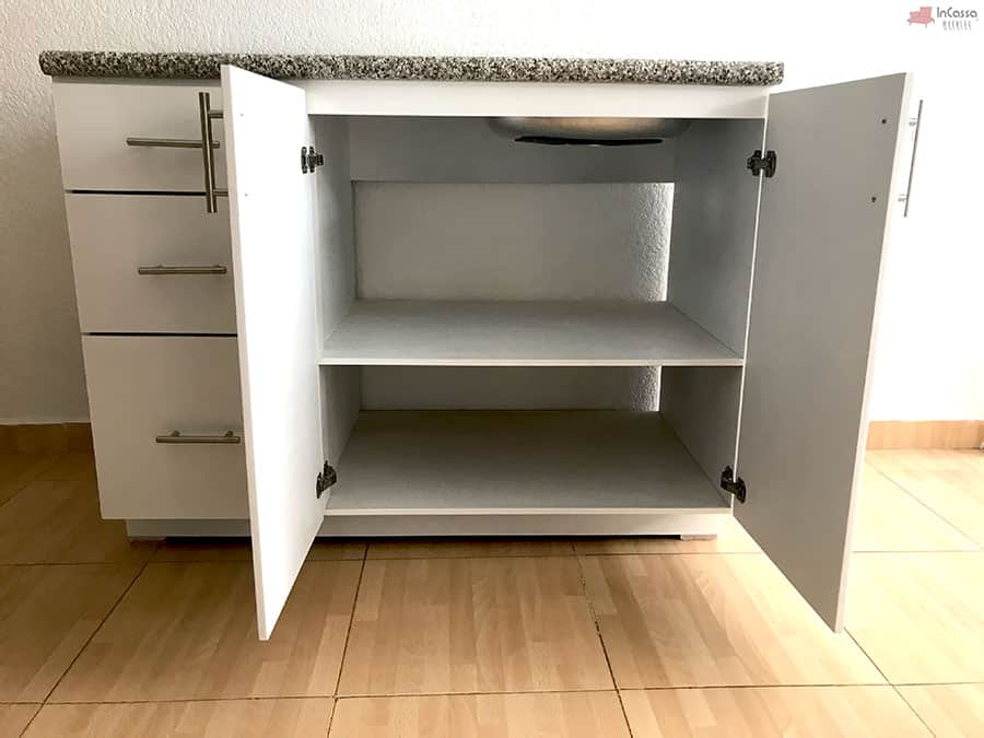 La imagen muestra de frente el módulo inferior de la Cocina Integral NEVADA de 2 metros, con ambas puertas abiertas para revelar dos amplios entrepaños en acabado blanco. Se aprecia el espacio destinado a la tarja en la parte superior, además de jaladeras de acero y una cubierta de formaica estilo granito purpina que aporta un toque moderno y elegante.