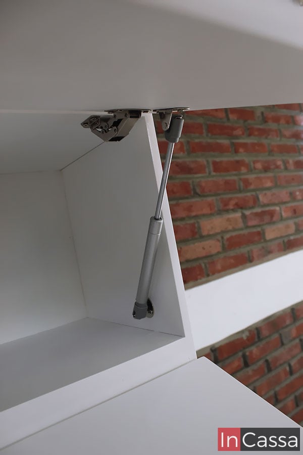 La foto se concentra en uno de los pistones neumáticos incorporados en las puertas del módulo sobre tarja, que en conjunto con bisagras metálicas hacen posible una apertura y cierre de manera vertical. La toma también permite apreciar parte del interior de la alacena, mismo que conserva el mismo tono blanco que se puede ver en las partes exteriores del mueble.