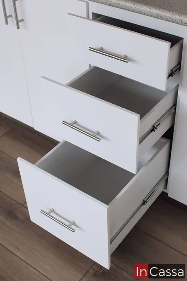 Una toma cercana de la sección con tres cajones del módulo inferior en la que, gracias a que se encuentran abiertos, podemos ver que su estructura completa mantiene el mismo color blanco presente en toda la cocina, También se nos enseña las diferentes capacidades interiores con las que cuenta cada cajón, además de su sistema de apertura y cierre guiado por rieles metálicos y sus jaladeras estilo T.
