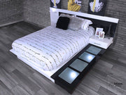 Cama moderna con cabecera con iluminación LED y burós integrados en tonos blancos y negros. Decorada con ropa de cama con diseño de escritura, cojines decorativos y detalles como una réplica de la Torre Eiffel y marcos de fotos. Ideal para un dormitorio elegante y contemporáneo, ofreciendo estilo y comodidad.