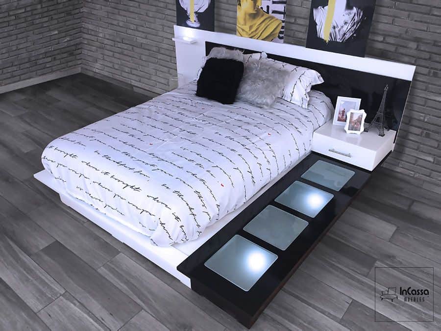 Cama moderna con cabecera con iluminación LED y burós integrados en tonos blancos y negros. Decorada con ropa de cama con diseño de escritura, cojines decorativos y detalles como una réplica de la Torre Eiffel y marcos de fotos. Ideal para un dormitorio elegante y contemporáneo, ofreciendo estilo y comodidad.