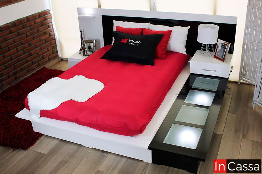 Recamara moderna con diseño minimalista, base con iluminación LED, cabecera y burós integrados en tonos blancos y negros. Incluye ropa de cama roja vibrante y cojines en blanco y rojo. Ideal para un dormitorio elegante y contemporáneo. Detalles decorativos como lámpara y marcos de fotos completan el ambiente.