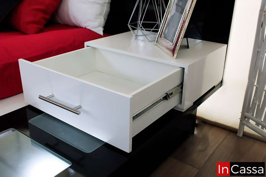 Buró blanco con cajón abierto, diseño moderno y elegante. Incluye manija metálica y detalles decorativos como un marco de fotos y una lámpara de diseño geométrico. Ideal para almacenamiento y decoración en un dormitorio contemporáneo.