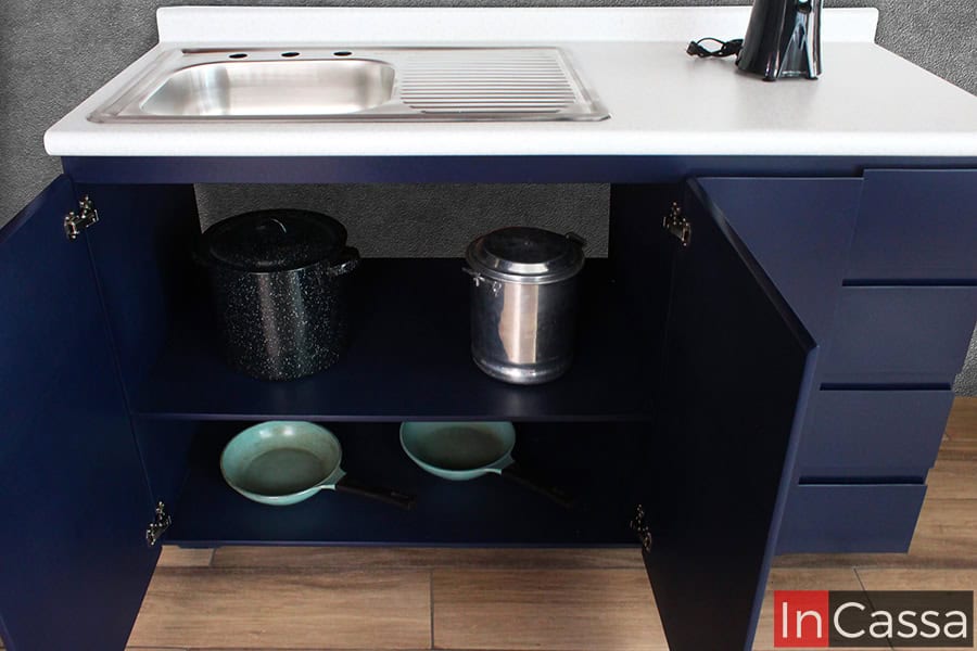 En esta imagen se observa el módulo inferior izquierdo de la cocina, equipado con una tarja de acero inoxidable sobre cubierta de formaica en estilo blanco fresh. Las puertas abiertas revelan dos estantes amplios para guardar ollas y sartenes. El diseño en MDF Chileno azul rey aporta elegancia y funcionalidad al espacio.
