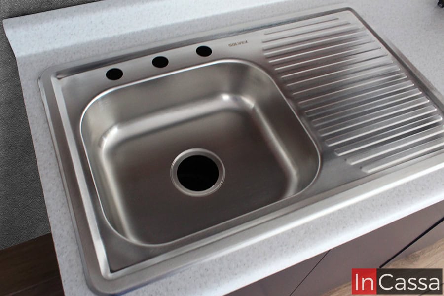 Una toma superior muestra la tarja de acero inoxidable con escurridor integrado, perfectamente montada sobre la cubierta de formaica blanco fresh de la cocina. El diseño incluye perforaciones listas para instalar mezcladora, dispensador y accesorios, ofreciendo practicidad y estilo en un solo elemento.