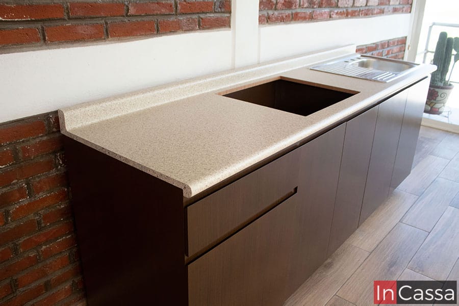 Una toma lateral del módulo inferior largo de la cocina, en la que se tiene una vista más completa de su estructura, la cuál muestra un acabado tabaco laqueado en su totalidad, y una cubierta de formaica en estilo sal y pimienta con tarja de acero inoxidable y un espacio para instalar una parrilla empotrable.