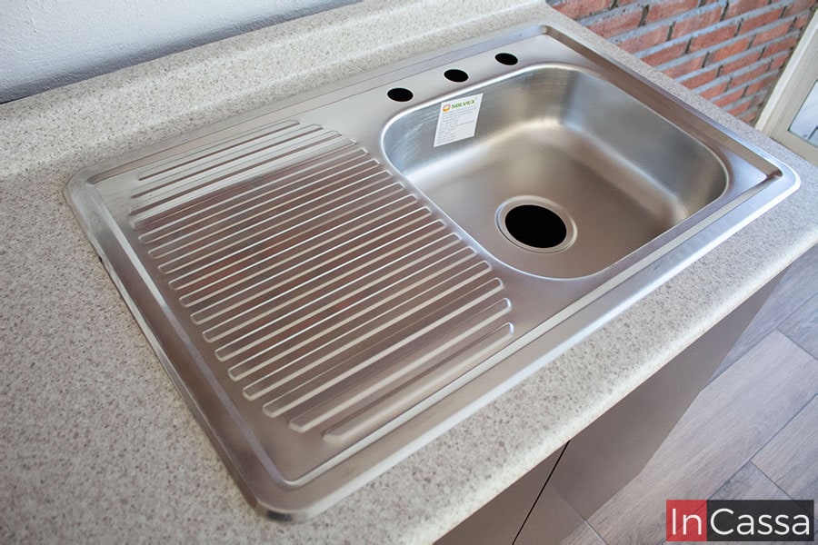 Está toma se enfoca en la tarja de acero inoxidable incluida en la cocina, está cuenta con una tina sencilla en su lado derecho y un escurridor en el izquierdo. Adicionalmente la foto también proporciona una vista parcial de la cubierta de formaica del módulo inferior, dejando apreciar su acabado en estilo sal y pimienta.