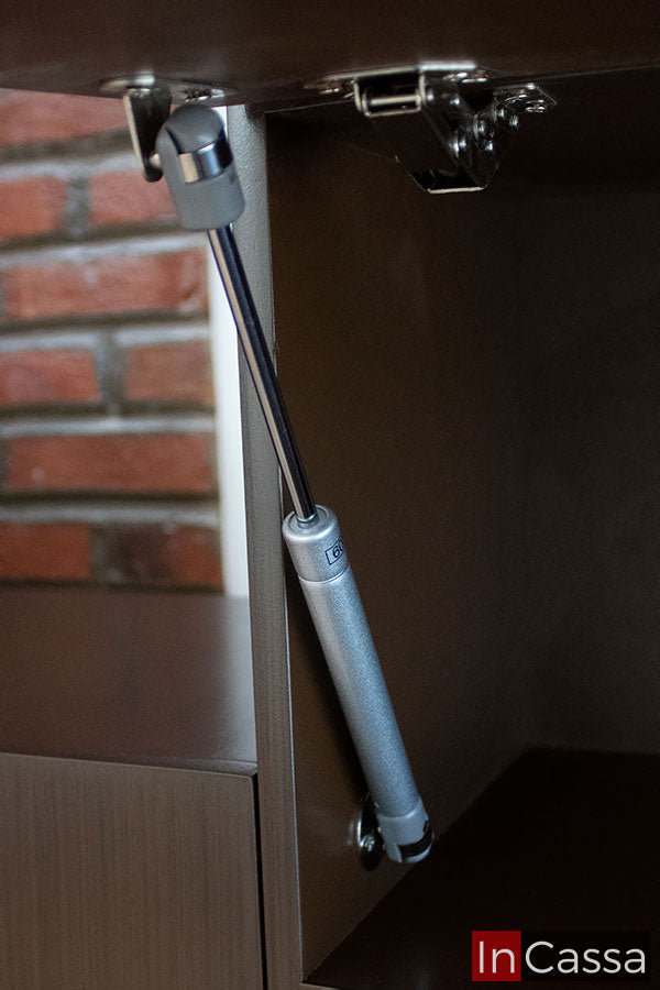 Una imagen de los pistones neumáticos integrados a las largas puertas de la alacena superior de la cocina, los cuales permiten su apertura y cierre de manera vertical. La toma también da un vistazo parcial al interior de la alacena, el cuál conserva el color café que puede verse en el resto de compartimentos.