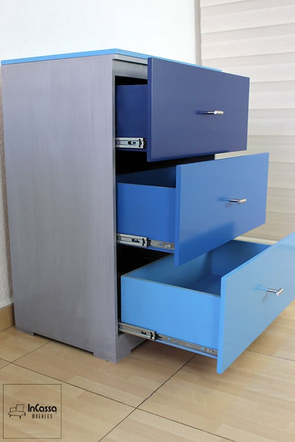 La imagen muestra un acercamiento lateral del cajonero incluido en la cama infantil, elaborado 100% en MDF chileno con acabado en tono gris cenizo claro. Los tres cajones presentan un diseño en tonos degradados de azul y están equipados con correderas metálicas de extensión que garantizan un deslizamiento suave. Las jaladeras metálicas cromadas complementan el diseño moderno y funcional.