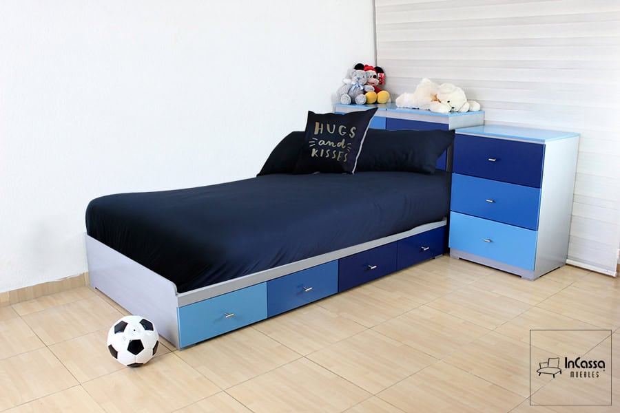La imagen presenta una cama individual en tonos azul celeste, azul medio y azul marino, con estructura en MDF acabado blanco cenizo. El diseño incluye seis cajones laterales con jaladeras metálicas, ideales para almacenamiento práctico. A juego, se observa un buró de tres cajones en los mismos tonos, reforzando el estilo juvenil y dinámico. Perfecta para habitaciones infantiles, combina funcionalidad y estética en un solo mueble.