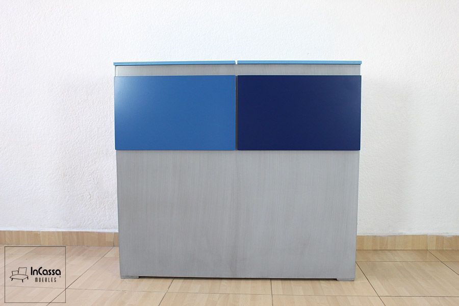 La toma muestra de frente el módulo respaldo con función de baúl del modelo Noa, fabricado en acabado gris cenizo claro. Está compuesto por dos tapas superiores abatibles en colores azul rey y azul marino, que aportan contraste visual. Su diseño limpio y compacto es ideal para aprovechar el espacio tras la cama, funcionando como respaldo y almacenamiento oculto al mismo tiempo.