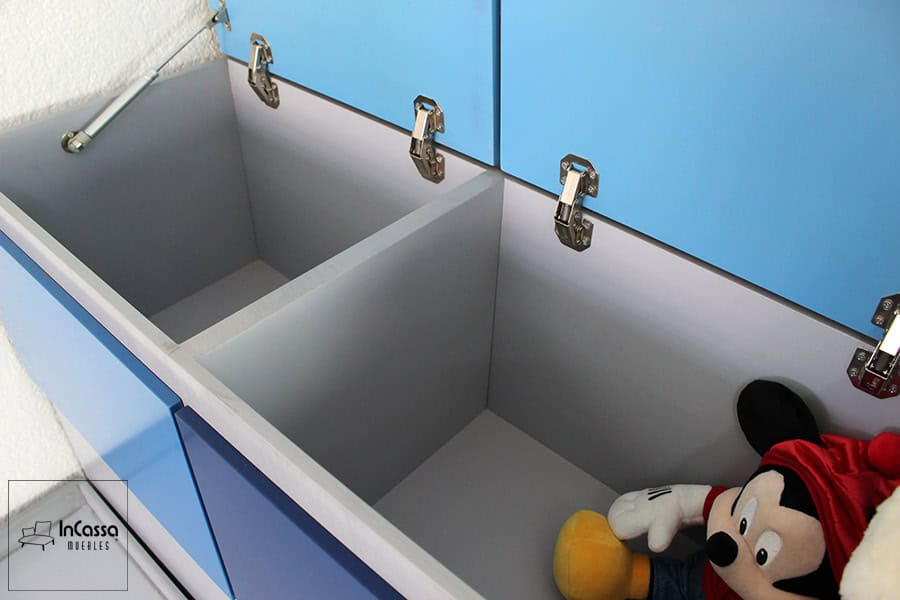 Se presenta una toma desde arriba que revela el baúl ubicado en la parte posterior de la cama infantil. El mueble, fabricado en MDF con acabado gris cenizo claro en el interior, cuenta con dos amplios compartimentos divididos, ideales para almacenar juguetes, ropa de cama o artículos personales. Las tapas en tonos azules están equipadas con bisagras metálicas resistentes y soporte de pistón neumático para una apertura suave y segura.