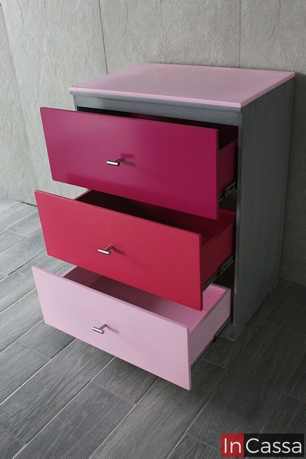 Esta imagen muestra un buró lateral infantil con tres cajones extendidos en una elegante composición de rosa pastel, rosa medio y fucsia. El cuerpo del mueble está fabricado en MDF chileno con acabado gris cenizo oscuro, mientras que la cubierta superior combina en rosa claro, ideal para habitaciones juveniles. Los cajones cuentan con correderas metálicas de extensión total y jaladeras cilíndricas cromadas, que aportan practicidad y un toque moderno al diseño.