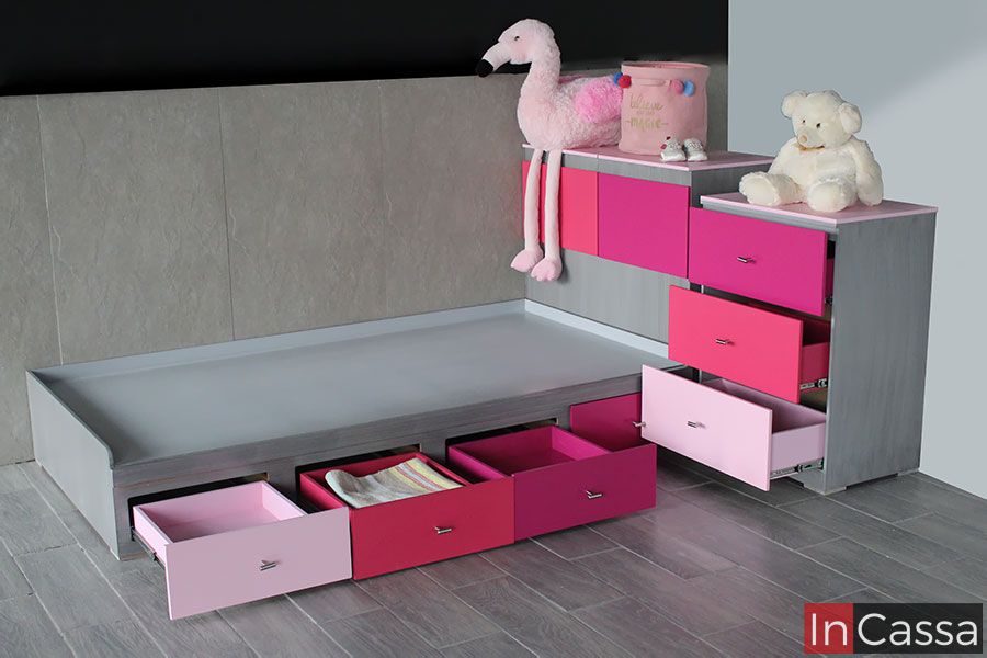La foto presenta una encantadora cama infantil en MDF chileno color gris cenizo oscuro con seis cajones inferiores completamente abiertos, pintados en una gama de tonos rosas degradados. A la derecha, un buró lateral y una cómoda alta con tres cajones más complementan el diseño. Las jaladeras metálicas cromadas y los rieles telescópicos aseguran un uso práctico y seguro. El acabado mate y los tonos vibrantes brindan un ambiente alegre y moderno ideal para niñas.