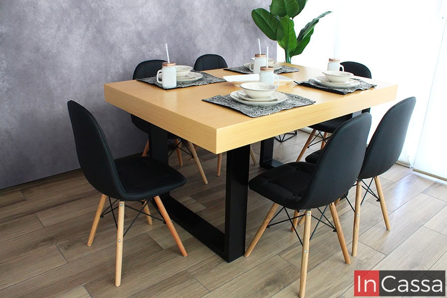 La imagen muestra un comedor para seis personas con mesa en acabado madera natural y base de MDF negra de forma rectangular Las sillas estilo eames combinan asientos negros con patas de madera clara. Un diseño moderno, luminoso y acogedor.