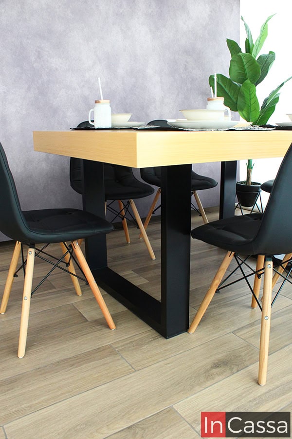 La imagen presenta un enfoque desde nivel bajo, destacando la robusta estructura de MDF en color negro que sostiene la mesa del Comedor NORMANDÍA. El contraste entre la base industrial y la cálida cubierta de madera clara define su estilo nórdico, acompañado de sillas eames negras con patas de madera, perfectas para un comedor moderno y sofisticado.