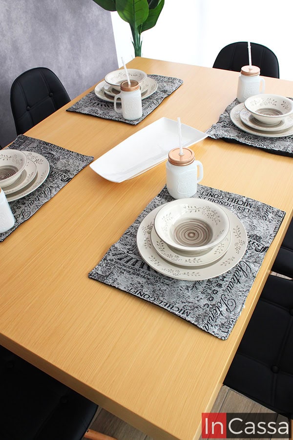 La fotografía ofrece una vista cenital que resalta la amplia cubierta de MDF en tono natural del Comedor NORMANDÍA. La disposición de la vajilla sobre individuales de tela en gris con estampado vintage añade un toque elegante y casual. Al fondo, las sillas eames negras con su distintivo diseño complementan este conjunto de estilo nórdico e industrial.