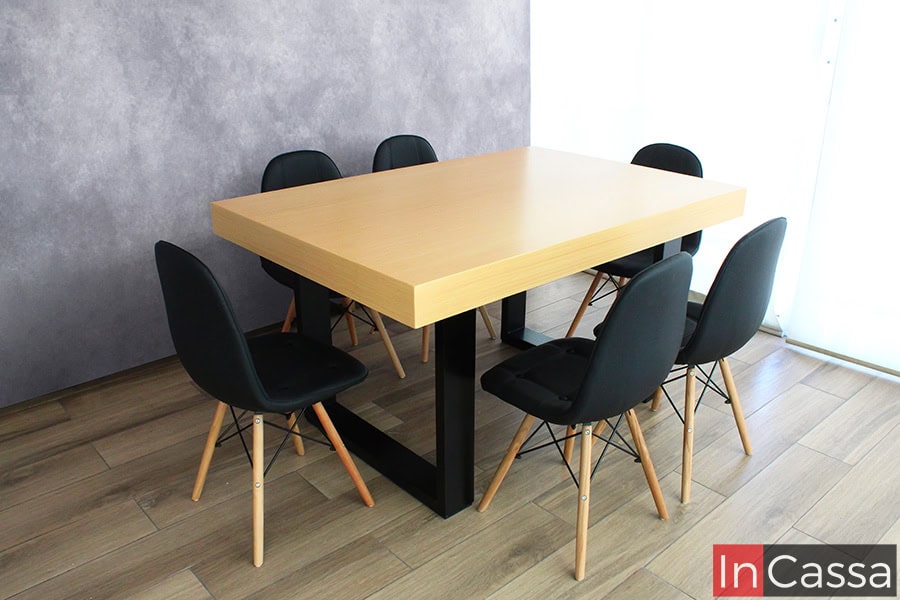 En esta imagen se aprecia el diseño limpio y robusto del Comedor NORMANDÍA en un ángulo lateral, donde resalta la gruesa cubierta de MDF en acabado natural. Las seis sillas tipo eames en color negro, con patas de madera natural y refuerzo metálico, rodean la mesa de manera simétrica, brindando un equilibrio perfecto entre estilo nórdico e industrial. El pedestal rectangular en tono negro completa la estética moderna del conjunto.