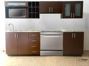 Una toma directa que presenta la distribución lineal de esta elegante cocina. Se observan seis módulos bien distribuidos: alacenas superiores con puertas lisas y de vidrio esmerilado, espacios abiertos para microondas y vinera, además de gabinetes inferiores con cubierta de formaica estilo granito purpina, tarja empotrada y espacio para estufa. Todo en un acabado color tabaco con jaladeras metálicas, ideal para un estilo moderno y funcional.