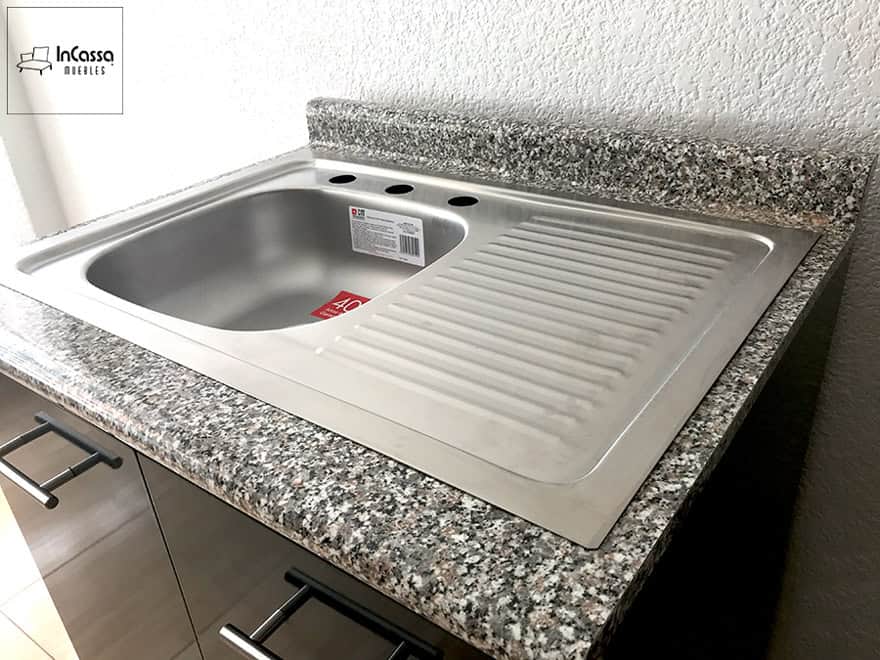 La imagen muestra un acercamiento lateral de la tarja de acero inoxidable con escurridor integrado, colocada sobre una cubierta de formaica tipo granito purpina. Se aprecian también las perforaciones listas para instalar mezcladora y accesorios.