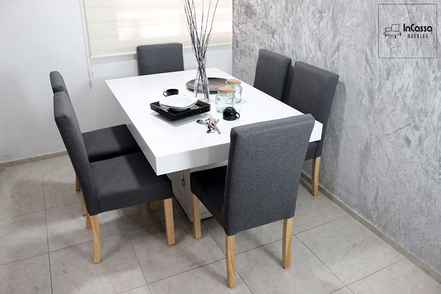Una fotografía de un comedor moderno que destaca por su mesa blanca de superficie lisa, acompañada de seis sillas tapizadas en pliana de un elegante tono gris cátala con patas de madera de pino. Este conjunto es perfecto para espacios más pequeños, manteniendo un estilo sofisticado y contemporáneo.