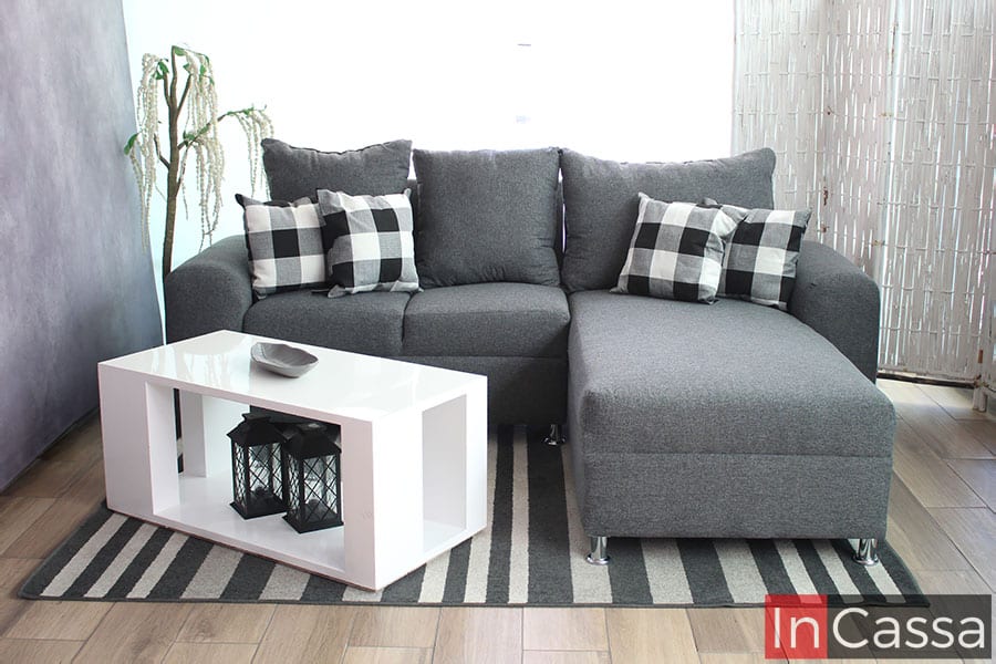 Sala minimalista en escuadra tapizada en lino mouse, decorada con 4 cojines pequeños y acompañada de una mesa de centro blanca. La sala es presentada en una estancia con piso de madera y una venta de pantalla completa.