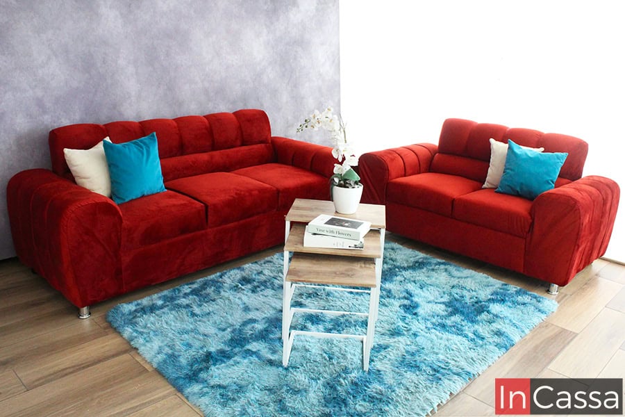 Fotografía en ángulo frontal que muestra un juego de sala Osaka en vibrante tapiz suede chilli, compuesto por sofá y love seat tapizados en textura suave. Destacan cojines decorativos en tonos crema y azul turquesa, colocados sobre una alfombra azul claro que aporta frescura. Al centro, una mesa auxiliar de madera y estructura blanca sostiene un arreglo floral, enmarcando la escena con un fondo lila y luz natural lateral.