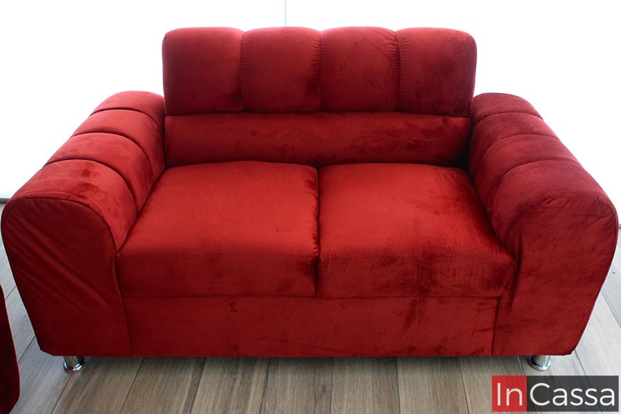 Imagen que muestra un love seat tapizado en aterciopelado tapiz suede color chilli, con costuras capitonadas en el respaldo y brazos acolchados de diseño envolvente. Las patas metálicas cromadas aportan un toque moderno, destacando sobre el piso laminado en tono madera clara.