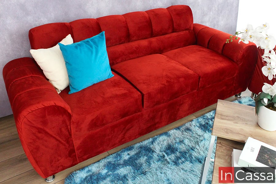 Imagen tomada en ángulo ligeramente elevado que resalta la textura aterciopelada del sofá de suede chilli, acompañado de cojines decorativos en tonos crema y azul turquesa. El mueble se ubica sobre una alfombra azul con textura mullida, junto a una mesa auxiliar de madera clara con estructura blanca, que sostiene un arreglo floral blanco, aportando frescura y contraste al conjunto.