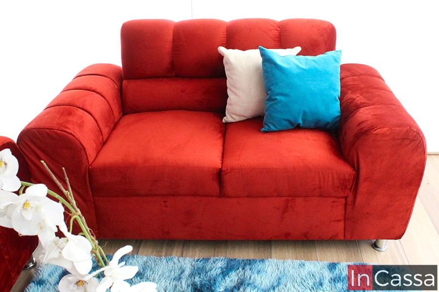 Toma frontal que destaca un love seat tapizado en suede chilli intenso con textura aterciopelada, complementado por cojines decorativos en color crema y azul turquesa. El mueble se ubica sobre una alfombra azul de textura suave, acompañado de un arreglo floral de orquídeas blancas que añade un toque elegante y fresco al espacio.