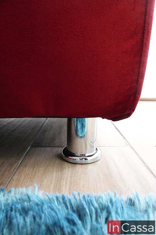 Toma en primer plano que resalta la pata cilíndrica metálica con acabado cromado, aportando estabilidad y un toque moderno al sofá tapizado en suede chilli. El reflejo de la alfombra azul y el piso laminado beige complementan la estética elegante y contemporánea del conjunto.