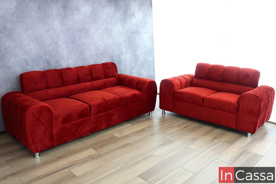 Fotografía frontal que presenta un juego de sala compuesto por sofá y love seat en vibrante tapiz suede chilli. El tapizado aterciopelado y las costuras capitonadas se combinan con patas metálicas cromadas, mientras el fondo gris y el piso laminado en tonos madera aportan un contraste elegante y moderno.