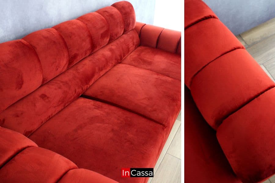 Se presenta un acercamiento al sofá de tres plazas en suede chilli, donde se aprecia la suavidad de la superficie y el mullido de los asientos. Las costuras capitonadas en respaldo y brazos realzan su diseño envolvente, resaltando sobre el piso laminado en tono madera clara.