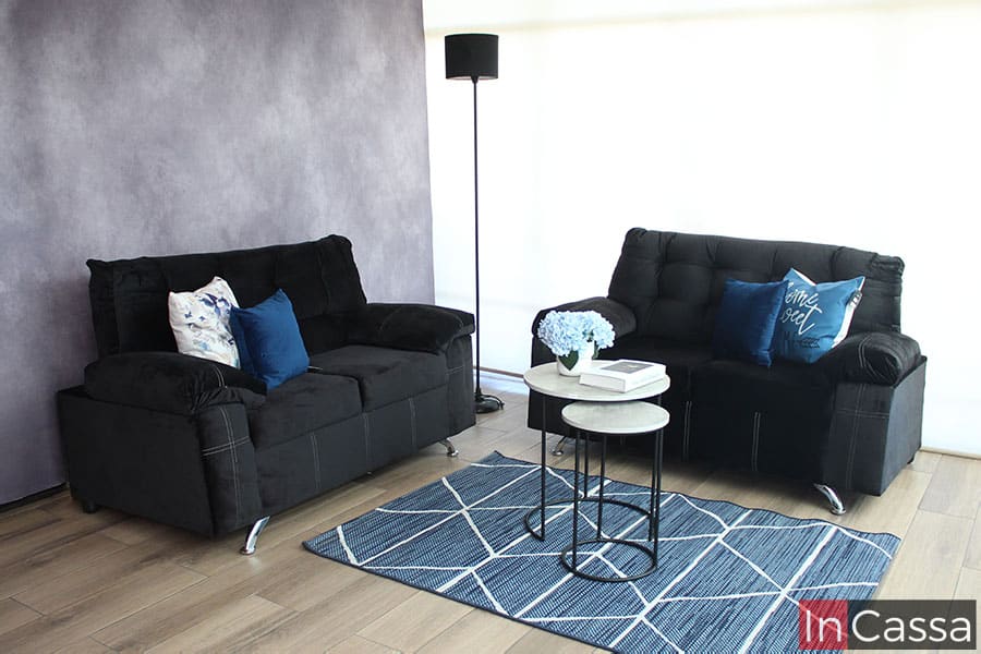 Sala 2 piezas tapizada en tela suede negro presentada en una estancia gris con piso de madera; la sala es adornada con 4 cojines de tonos azulados y turquesa.