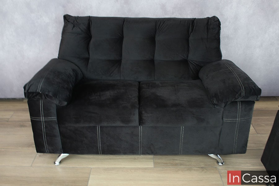 La fotografía presenta uno de los love seat en una vista de frente acomodado en una pared gris, donde podemos observar su tapizado en suede negro completo, además de las patas metálicas delanteras con acabado cromado que resaltan en el piso de madera.