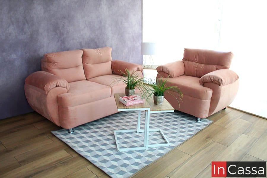 La imagen presenta la Sala Modelo PAMPLONA tapizada en suede color rosa palo, conformada por un love seat y un sillón. El ambiente moderno con muro lila y piso de madera resalta su diseño acolchado, suaves líneas curvas y patas metálicas cromadas.