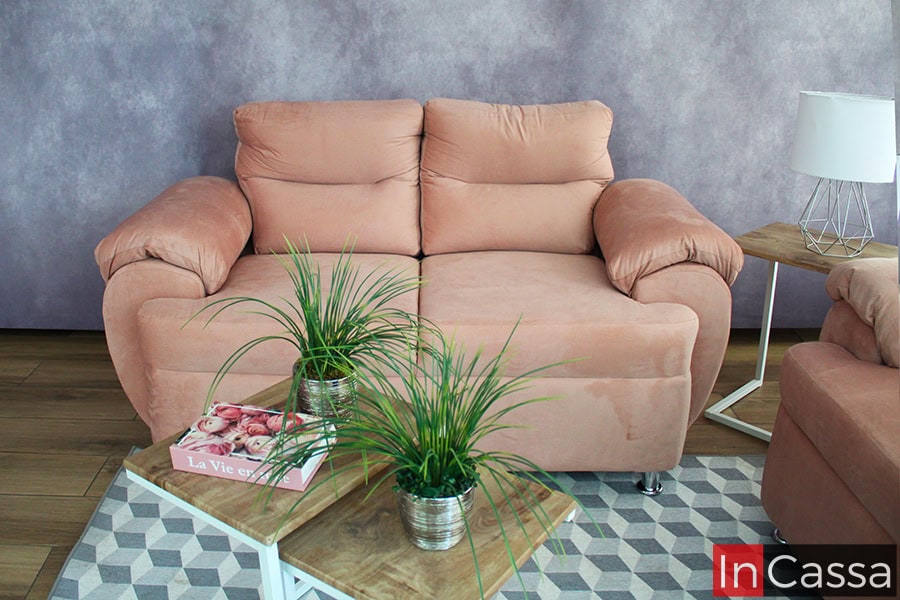 En esta toma se aprecia el love seat de la Sala Modelo PAMPLONA, tapizado en suede rosa palo. Su diseño acolchado, brazos amplios y patas metálicas cromadas aportan comodidad y elegancia. Fondo con muro lila y mesa lateral con lámpara decorativa.