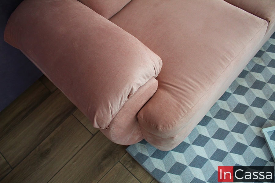 La imagen muestra un acercamiento del brazo acolchado del love seat PAMPLONA, tapizado en suede rosa palo. Se aprecia su textura suave y costuras finas sobre el tapete geométrico, destacando la comodidad y el acabado elegante del diseño.