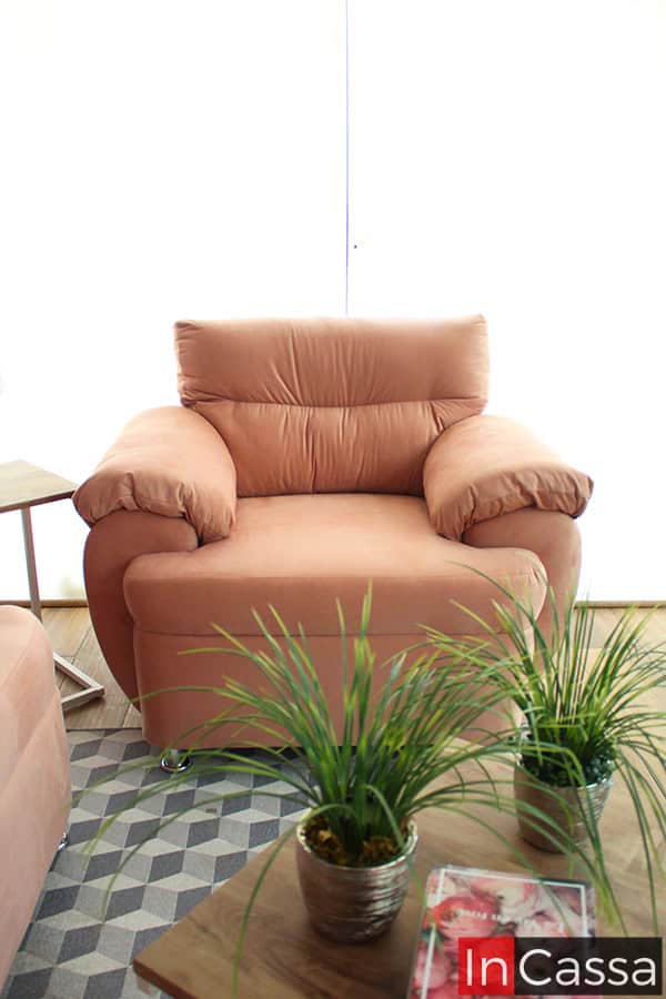 La toma presenta el sillón individual de la Sala Modelo PAMPLONA, tapizado en suede rosa palo. Su estructura robusta y brazos anchos reflejan confort y elegancia, complementado con patas cromadas y fondo iluminado que realza su tono suave.