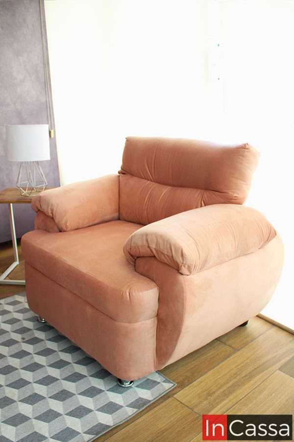 La imagen muestra el sillón individual del modelo PAMPLONA en vista lateral, tapizado en suede rosa palo. Destaca su respaldo mullido, brazos redondeados y costuras delicadas sobre piso de madera y tapete geométrico, creando un ambiente moderno y acogedor.