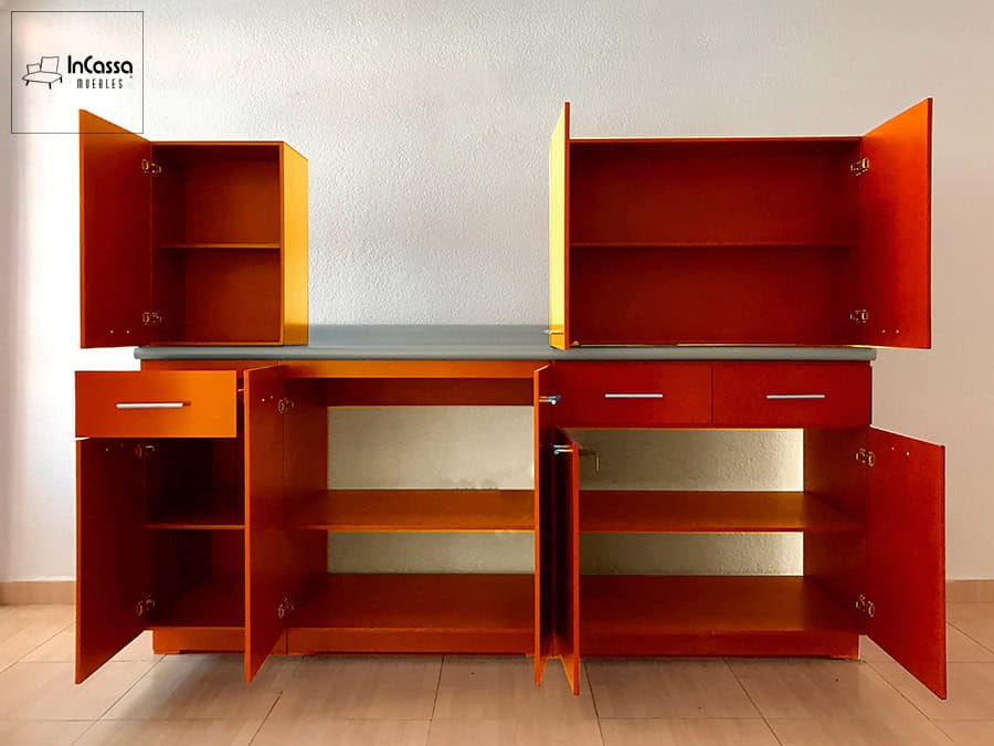 La imagen muestra la cocina PHOENIX para Parrilla en color mandarina con todos sus módulos abiertos, revelando la amplitud de almacenamiento tanto en alacenas superiores como en gabinetes inferiores. Destaca la cubierta de formaica en tono aluminio que contrasta elegantemente con el acabado vibrante.