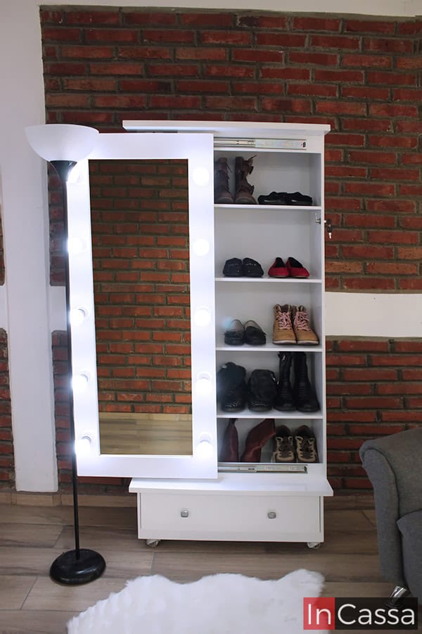 En esta toma frontal se aprecia la zapatera Piacenza en color blanco, con espejo lateral iluminado por diez focos LED. A la derecha, cinco repisas internas exhiben distintos pares de calzado y en la parte inferior se integra un cajón con jaladeras circulares. El fondo de ladrillo realza su diseño moderno.