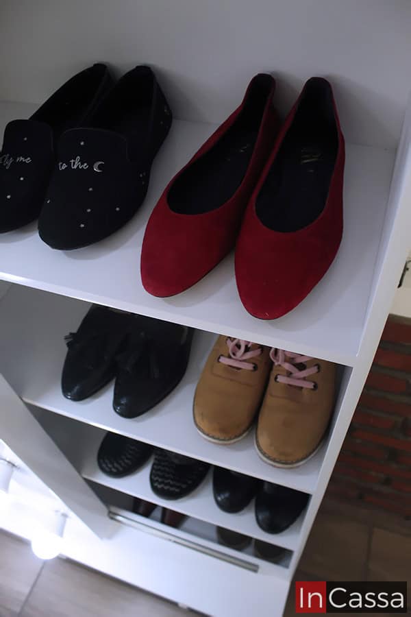 La imagen presenta un acercamiento lateral de las repisas internas de la zapatera Piacenza blanca, mostrando pares de calzado en diversos estilos y colores, como mocasines negros, flats rojos, botines beige y zapatos formales negros. La iluminación LED del espejo aporta un toque elegante y funcional.