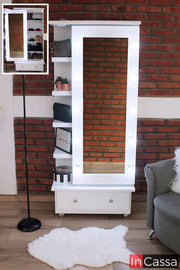 La foto presenta una toma frontal de la zapatera Piacenza en color blanco, con espejo de cuerpo completo enmarcado por bombillas LED encendidas. A la izquierda destacan sus repisas laterales internas con decoración y a la base un cajón amplio, todo sobre ruedas para fácil movilidad.